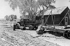 Gentleman standing next to old pickup truck on the right, parked in front of wooden barn-like house, trees, flat bed trailer and pieces of  wood and barrels are scattered about.