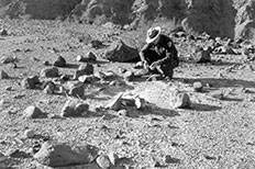 Park ranger in uniform squats to the ground with his head down as he holds items from the campsite,  he is surrounded by different sized rocks and desert landscape.