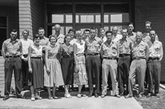 A group of five women and fourteen men posing for a photo in front of brick building with windows.

