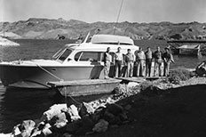 Seven men pose together in front of docked boat surrounded by rocky textured mountains.
