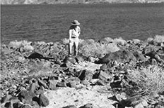 Man standing in front of Lake wearing a hat with his left leg perched on a rock overlooking a campsite fire ring.