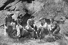Park ranger in uniform gestures with hands while four people sit on rocks with him, petroglyphs on rocks behind them.