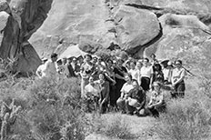 Group of men and women posing in front of rock formations with petroglyphs. 