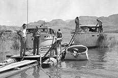 Two men pose together on the dock while three other men attend to three boats that float on the river.