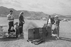 A woman roasts hot dog at a brick barbeque on the beach, five others are near her,10 cars parked in distance.