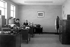 A brick office with two windows and three desks at left, filing cabinets to the right, two women work at desks.