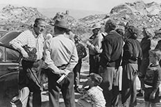 Gentleman with microphone and headphones leans on car door as he talks to a man in uniform with his hands behind his back as a group of onlookers listen. 