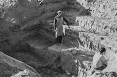 Older man in hat and overalls with shovel nearby standing in a hole with another man in the left corner with his back towards the camera. 