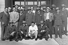 Group of men in suits and NPS uniforms pose for a picture in front of brick building with windows. 

