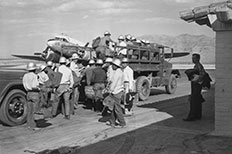 Group of men wearing hard hats boarding on a large truck carrying equipment bags, a prop plane is in the background on the left. 