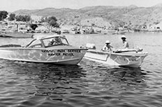 A man in a National Park Service Ranger Patrol boat floats on the Lake next to two men wearing pith helmets in a smaller boat.