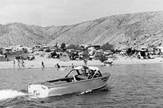 Park Ranger steering patrol boat on the cove as people swim near the shore, parked cars and tents are scattered across the sand. 