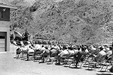 Gentleman giving speech on stage next to seated marching band, American flags displayed and crowd of people listen while sitting in folding chairs. 