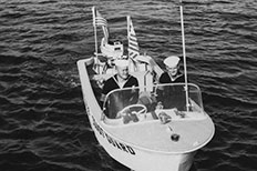 Two uniformed U.S. Coast Guard men steer their boat in water with two American flags on the back and U.S. Coast Guard painted on the side.