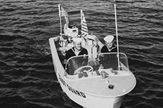 Two uniformed U.S. Coast Guard men steer their boat in water with two American flags on the back and U.S. Coast Guard painted on the side.