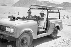 Two lifeguards take a drive on the beach in door-less jeep as beach goers swim in the water.