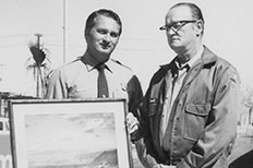 Two men in NPS uniforms pose next to each other as one holds framed artwork.