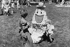 Tommy Trout a puppet-like mascot sitting down in grass entertaining two small children. 