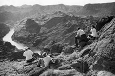 Four hikers sit on rock formations and observe their view of the river far below which is surrounded by towering rock. 