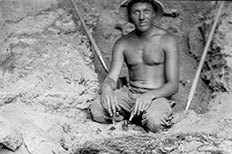 Man wearing hat and holding tools in his hands while sitting in front of a cave with a fire pit made of wood and twigs displayed before him.