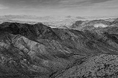 View of ridged and rocky rolling mountains with desert plants scattered across and a small glimpse of a body of water to the right-hand side. 