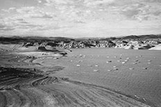 Shoreline with tire imprints and a body of water to the right with boats scattered across, surrounded by mountains.