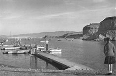 Girl in pigtails stands on beach beside lake dock with boats tied up to it.