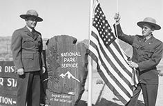 One Ranger in uniform stands next to a National Park Service sign while another raises the American Flag on a flagpole.