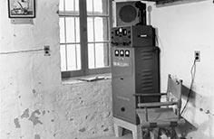 Communication equipment in corner of a room by a window with a folding chair next to it.