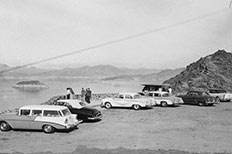 Six cars parked side by side along a stone  wall and people observe a body of water surrounded by mountains. 