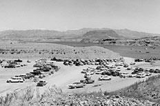 Many cars parked on the shoreline surrounded by desert landscape and a body of water with boats scattered across it, mountains in the distance.