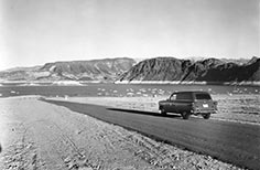 Car driving on road that goes towards a lake with mountains in the background.