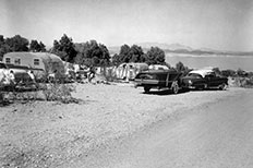 A clothesline hung between two parked camping trailers and surrounded by other parked cars and trees, a view of lake and mountains in the distance.