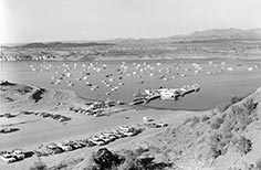 Many cars in parking lot nearby lake with a dock and many boats on it.