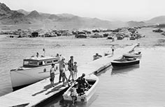 People stand on a dock with several boats tied up to it and many cars parked in distance.