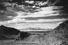 Muddy and textured mountains with a glimpse of a body of water behind them and mountains in the distance with a cloudy sky above. 