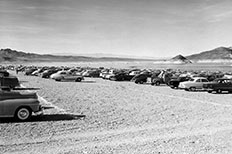 Rows of parked cars side-by-side on the shoreline overlooking lake, mountains in distance.