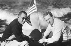 Two men sit in a boat on a lake with an American flag attached.