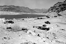 Mountains with a body of water in distance, parked cars and tents along the shoreline. 