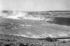 Desert landscape with tire markings across the flat plain as clouds of dust cover the mountains in the distance.