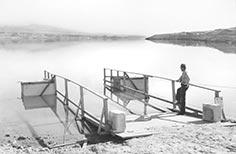 Man in uniform stands next to boat launch on a lake.