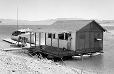 Man leans against floating building attached to dock on a lake with boat tied up to it.