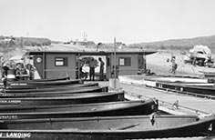 People stand under structure at end of a dock on a lake with many boats tied up to it.
