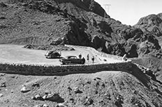 Textured mountains with a dirt parking area below that is surrounded by a stone wall. Two cars four people.