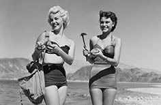 Two women in swimsuits stand holding rocks, one with a bag on her shoulder and another with a hammer.