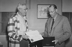 Two men stand each holding one side of an award and one holding a portfolio.