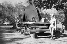 Woman sits at a picnic table in front of a tent with two children nearby.