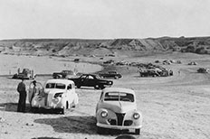 Two parked cars facing the camera foreground, many cars and lake shoreline behind.