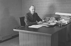 Man sits at a desk with papers and a telephone on it.