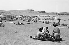 Many parked cars and crowds of people on a beach by a lake.